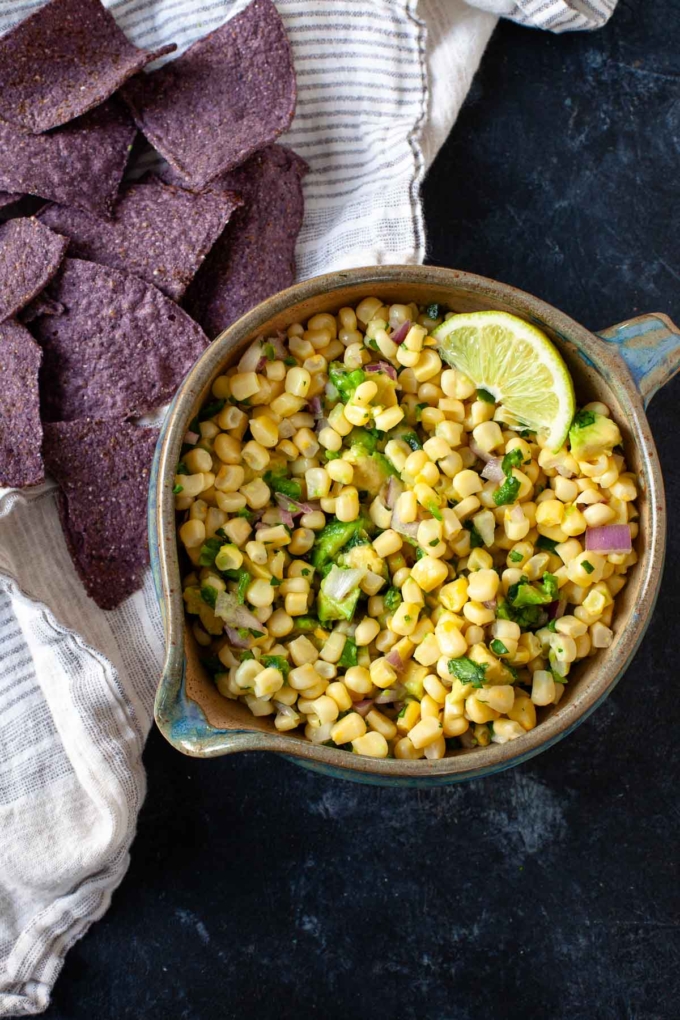 corn avocado salsa with lime wedge in brown and blue ceramic bowl next to blue tortilla chips on dish towel