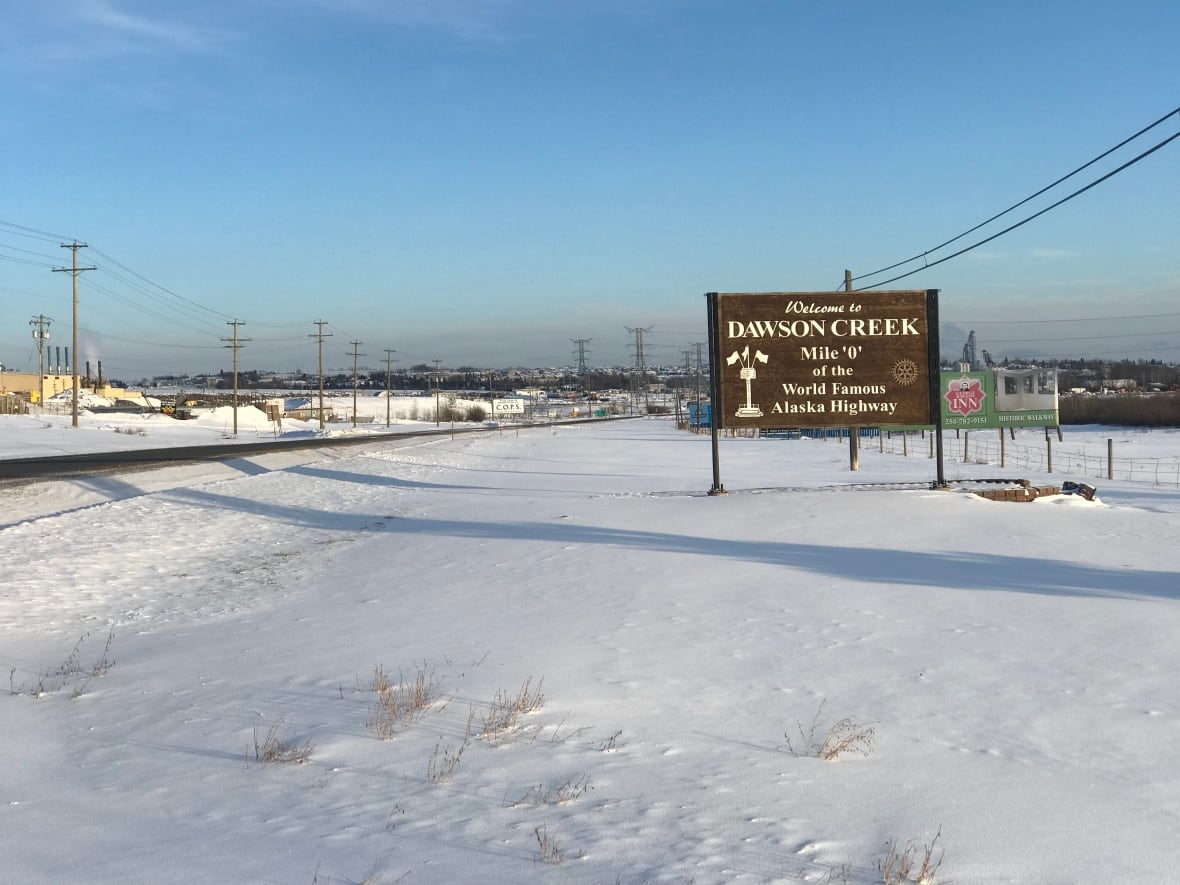 A wooden sign at the entrance to a snowy community reads, 'Welcome to Dawson Creek, Mile 0 of the World Famous Alaska Highway.'