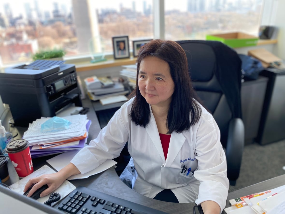 An Asian doctor wearing a lab coat works at a computer in her office.