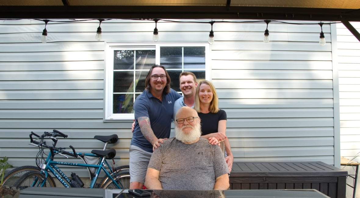 Three smiling people put their arms and stand behind a seated man, who is also smiling.