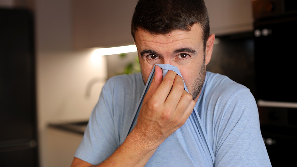 Man covering his nose with his shirt, looking into camera, after smelling something bad.